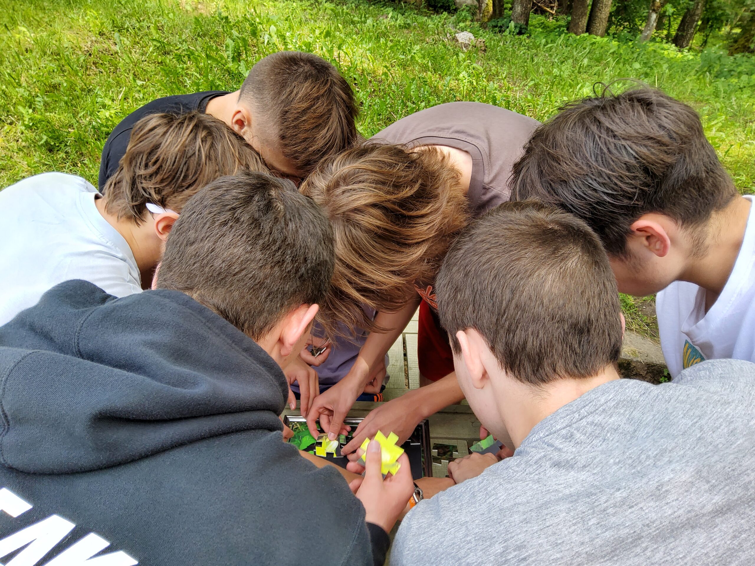 Group of young boys gathered around a table, engaged in a collaborative activity outdoors, surrounded by greenery. They are focused on assembling or playing with colorful toys or games.