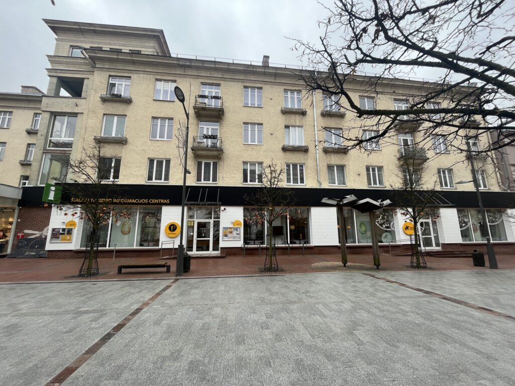 Facade of a yellow apartment building with balconies, featuring storefronts on the ground level and a paved plaza in front, under a cloudy sky.