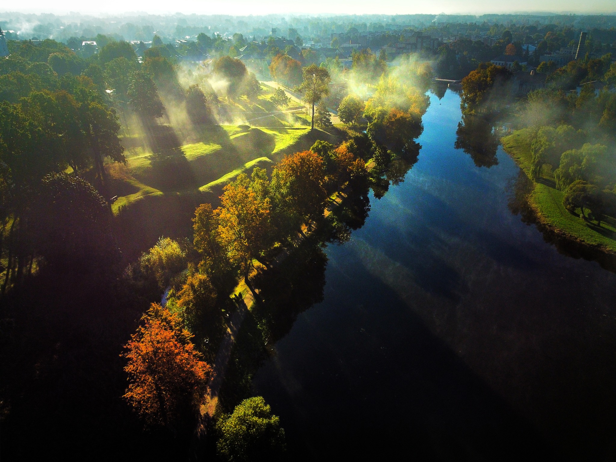 Aerial view of a serene river surrounded by lush greenery and autumn-colored trees, with morning mist hovering over the water and sunlight casting shadows on the landscape. Aerial view of a serene river surrounded by lush greenery and autumn-colored trees, with morning mist hovering over the water and sunlight casting shadows on the landscape.
