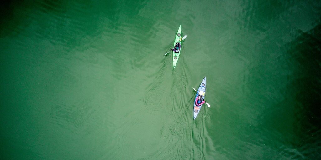Two kayakers paddling in tranquil green waters, viewed from above, showcasing outdoor adventure and water sports.