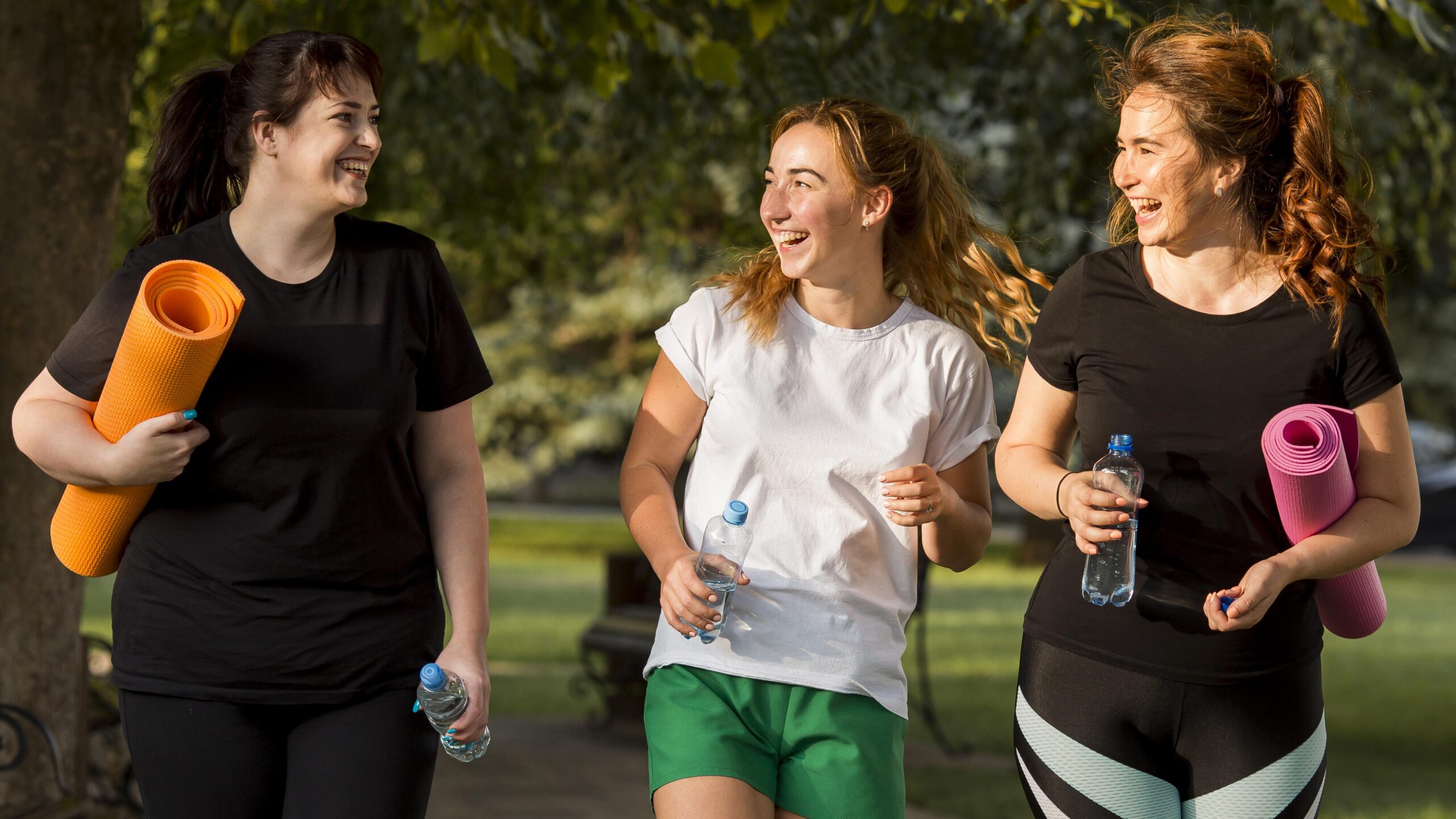 Three women jogging together in a park, smiling and holding water bottles, promoting fitness and friendship.