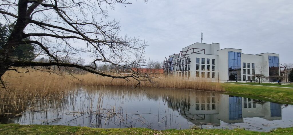 Modern building reflected in a calm pond surrounded by tall grasses and bare trees under a cloudy sky. Modern building reflected in a calm pond surrounded by tall grasses and bare trees under a cloudy sky.