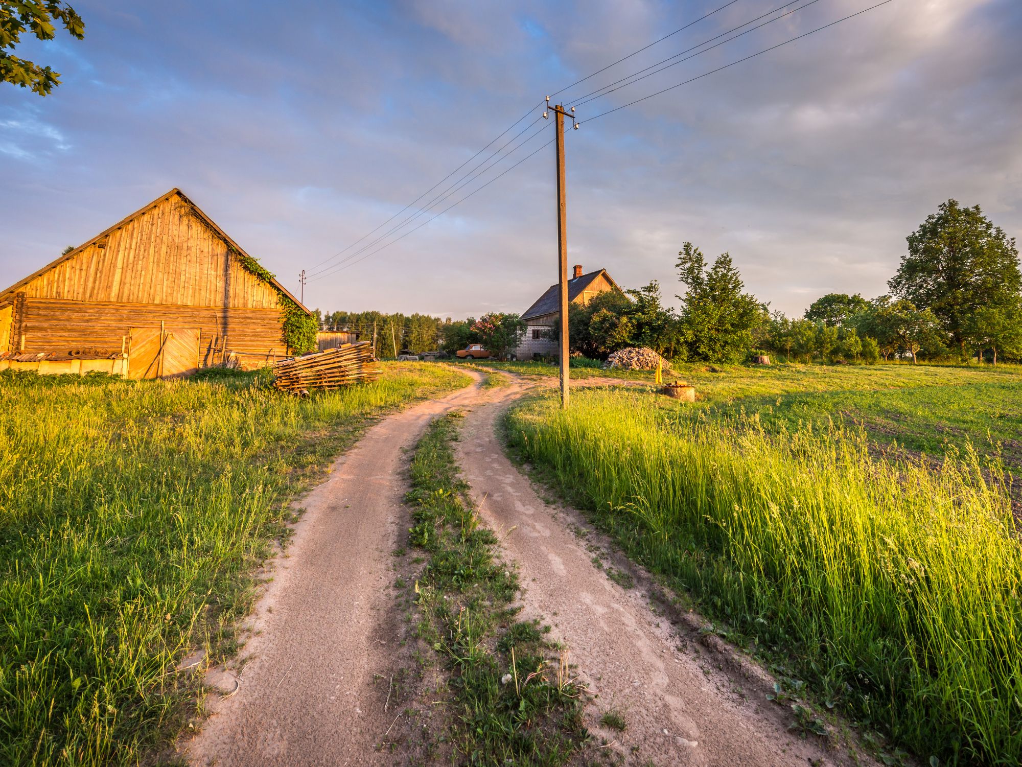 Country road leading through lush green fields with wooden houses under a clear sky, depicting a serene rural landscape. Country road leading through lush green fields with wooden houses under a clear sky, depicting a serene rural landscape.
